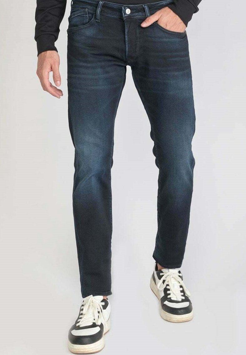 Personne portant un jean slim bleu foncé et des baskets noires et blanches, une main dans la poche et l'autre détendue sur le côté.