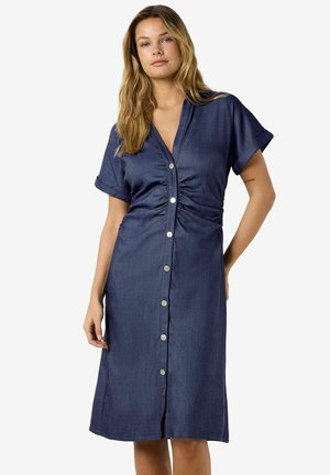 Naine, kes kannab põlveni ulatuvat tumesinist nööpidega kleiti, millel on lühikesed rullitud varrukad ja vöökoha peal kortsud.