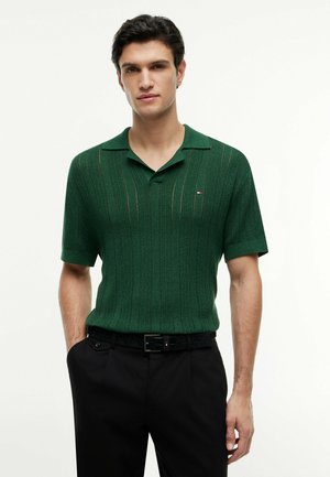 Mężczyzna w ciemnozielonym, krótkim, dzianinowym polo z subtelnymi pionowymi paskami i czarnych spodniach, stojący z jedną ręką w kieszeni.