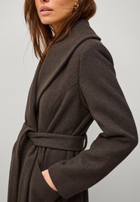 Cappotto avvolgente marrone, texturizzato, con un ampio colletto, cintato in vita, dotato di tasche profonde e una vestibilità sartoriale. Materiale morbido simile alla lana.