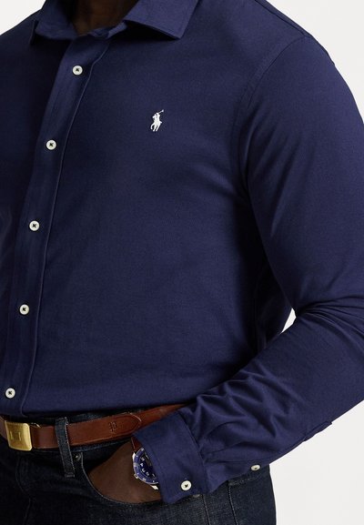 Ναυτική πουκάμισο με κουμπιά σε εφαρμοστή γραμμή, φτιαγμένο από ομαλό ύφασμα. Διαθέτει μικρό κεντητό λογότυπο αλόγου, τσέπη στο στήθος και λευκά κουμπιά.