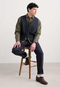 Chaleco de mezclilla oscuro sobre una camisa a cuadros verdes, combinado con pantalones oscuros y zapatos de cuero marrón, sentado en un taburete de madera.