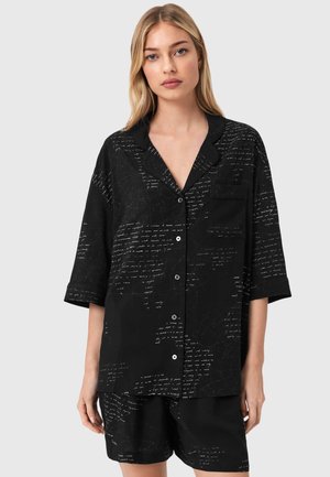 Vrouw met blond haar die een loszittend zwart overhemd met knopen draagt en bijpassende korte broek met wit schriftpatroon op lichtgrijze achtergrond.