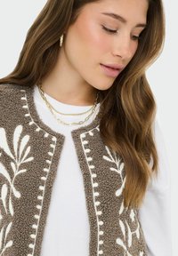 Jeune femme aux longs cheveux bruns portant des boucles d'oreilles créoles dorées, des colliers en or superposés, un haut blanc et un gilet marron texturé avec des motifs floraux blancs.