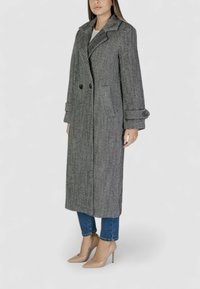 Cappotto a scacchi grigio e bianco con colletto a punta, dotato di bottoni doppi e due tasche laterali. Indossato sopra jeans blu e tacchi nudi.
