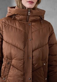 Giacca a vento marrone con colletto alto, dotata di tasche con zip e bottoni a pressione. Design trapuntato e testurizzato con una finitura calda.