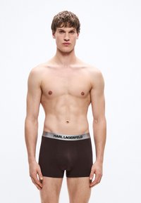 Calzoncillos bóxer marrones con una cintura plateada que muestra "KARL LAGERFELD." Tela suave y diseño ajustado para mayor comodidad. El modelo no lleva camiseta.