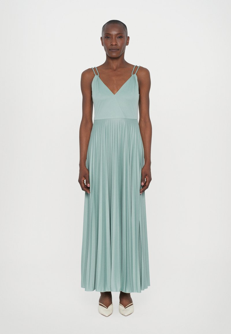 Robe maxi vert clair avec décolleté en V, design plissé et deux fines bretelles. Le tissu est lisse, associé à des chaussures blanches pointues.