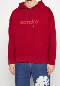 Czerwony sweter z kapturem, z przednią kieszenią, sznurkiem do ściągania i dużym logo w jaśniejszym odcieniu. Wykonany z miękkiego materiału z prążkowanymi mankietami.
