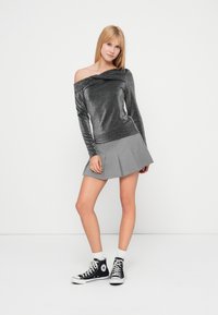 Γκρι μεταλλικό τοπ με μακριά μανίκια και ώμους ακάλυπτους, σε συνδυασμό με πλισέ γκρι φούστα και μαύρα sneakers ψηλά στον αστράγαλο, τοποθετημένα σε λευκό φόντο.