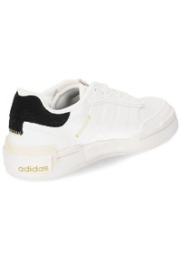 White sneaker with a black suede accent at the heel, gold detailing, and a textured white sole. Features three white stripes on the side.