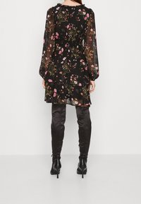 Nainen, joka on pukeutunut läpinäkyvään mustaan kukkamekkoon, jossa on pitkät hihat ja kerroksellinen hameosa, yhdistettynä mustiin reittä yltäviä korkokenkiä, kuvattu takaa.