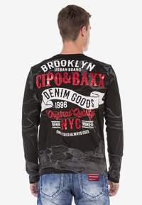 Schwarzes Langarmshirt mit großen, kräftigen Textgrafiken in Weiß und Rot, mit einem abgenutzten Textur und Markenelementen auf der Rückseite.