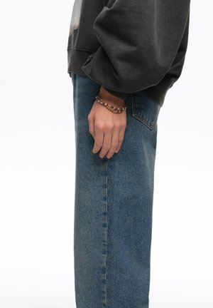 Persona vestida con vaqueros azules y una camiseta de manga larga oscura, con una pulsera de cuentas en la muñeca izquierda, de pie contra un fondo liso.