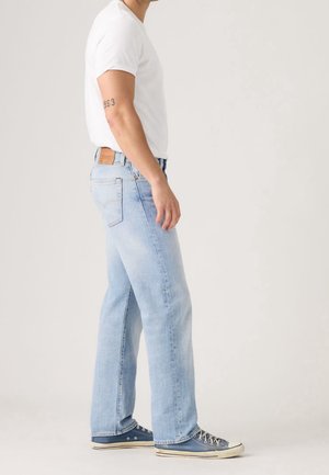 Man die lichtblauwe spijkerbroek, wit T-shirt en blauwe sneakers draagt, staand zijwaarts op een effen achtergrond.