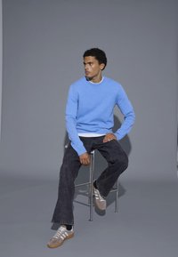 Világoskék, kerek nyakú pulóver, kötött textúrával, fekete farmernadrággal és fehér csíkos szürke tornacipővel párosítva, egy bárszéken ülve.