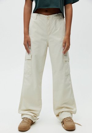 Pantalons cargo beiges à coupe ample, dotés de plusieurs poches latérales, d'une fermeture à bouton et d'un tissu en coton texturé. Associés à des baskets marron clair.