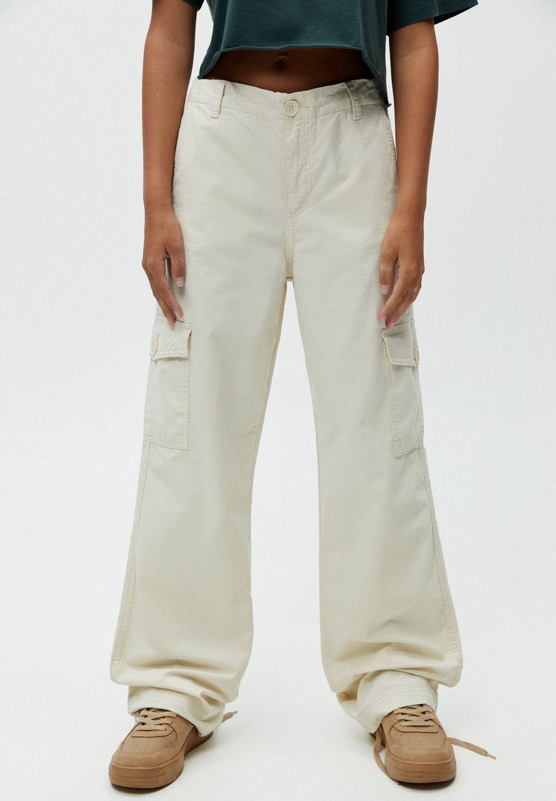 Beige cargo byxor med lös passform, med flera sidofickor, knappstängning och en texturerad bomullstyg. Kombinerat med ljusbruna sneakers.