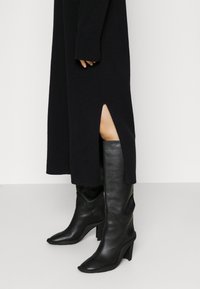 Schwarze knielange Lederstiefel mit spitzer Zehe und blockartigem Absatz, kombiniert mit einem langen schwarzen Strickkleid mit Seitenschlitzen.