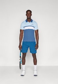 Blå polo skjorta med horisontella ränder i ljusblått och vitt, parat med matchande blå shorts. Håller en tennisracket och bär vita sneakers.