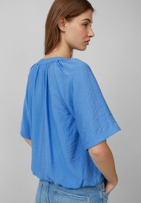 Top bluetto a maniche corte con una finitura texturizzata e stropicciata, caratterizzato da un collo arricciato e una vestibilità morbida, abbinato a jeans in denim chiaro.