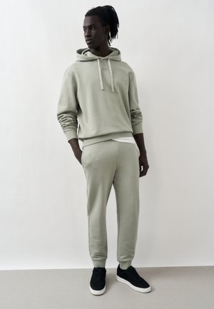 Bărbat cu dreadlocks purtând hanorac gri deschis și pantaloni de trening asortați, stând cu mâinile în buzunare, uitându-se spre stânga pe un fundal simplu.