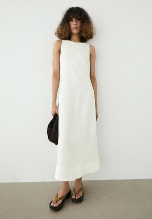 Giovane donna con capelli ricci che indossa un vestito midi bianco senza maniche e sandali con plateau, che tiene una piccola borsa nera davanti a uno sfondo semplice.