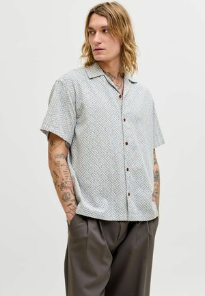Jeune homme aux longs cheveux et tatouages, portant une chemise à manches courtes à motifs boutonnée et un pantalon ample de couleur foncée, les mains dans les poches, regardant vers la gauche.