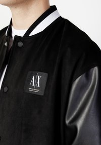 Svart bomberjacka med en läderliknande kropp och ärmar i läder, med en vit randig ribbad krage och en A|X-logotypapplikation på framsidan.