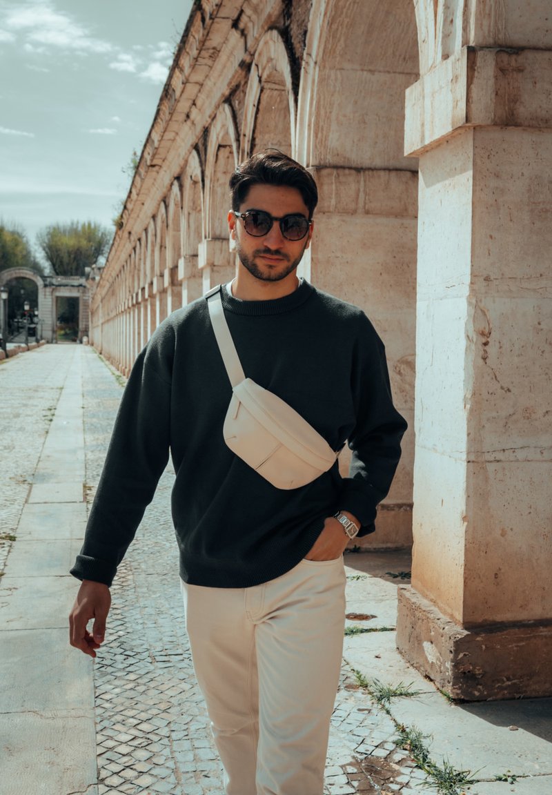 Muž v tmavém svetru a bílých kalhotách kráčí po kamenné cestě. Nosí sluneční brýle a lehkou krossbody tašku. V pozadí jsou klenuté kamenné struktury.