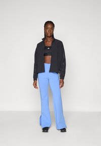 Črna kratka zip jakna nad črnim športnim modrčkom, v kombinaciji z svetlo modrimi širokimi hlačami. Gladek material z atletskim krojem in logotipnimi poudarki.