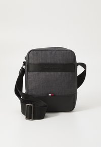 Gray fabric crossbody bag with black accents, featuring a logo strap, zip closure, and a small red, white, and blue detail at the bottom.