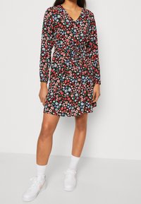 Blumenkleid mit schwarzem Hintergrund, das rote, blaue und grüne Blumen zeigt; mit langen Ärmeln, Wickel-V-Ausschnitt und verstellbarem Taillenzug. Kombiniert mit weißen Sneakers.