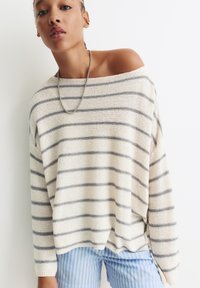 Jeune personne portant un pull crème à épaule dénudée avec des rayures grises, un collier en chaîne argenté et un pantalon rayé bleu et blanc, sur fond blanc.