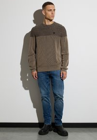 Brauner Strickpullover mit strukturiertem Oberteil, Rundhalsausschnitt und gerippten Bündchen, kombiniert mit blauen Jeans und schwarzen klobigen Stiefeln.