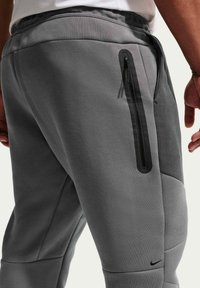Nærbillede af person iført grå atletikbukser med sort lynlåslomme og lille sort Nike-swoosh på låret.