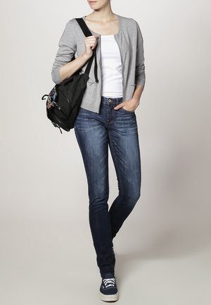 Kobieta ubrana w szary kardigan, biały top, niebieskie dżinsy, trampki, niosąca czarny plecak na jednym ramieniu, stojąca na tle jednobarwnym.