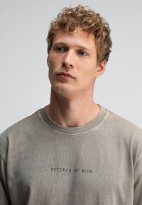 Graues Crewneck-Sweatshirt aus weichem Material, mit dunklem Druck "BUTCHER OF BLUE" auf der Vorderseite sowie gerippten Bündchen und Kragen.