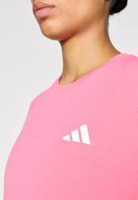 Ροζ βαμβακερό μπλουζάκι με στρογγυλή λαιμόκοψη, κοντά μανίκια και λευκό λογότυπο με τρία στοίβας τρίγωνα στη αριστερή πλευρά.