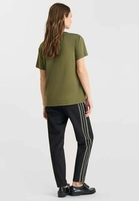 Olivengroene shirt met korte mouwen, gecombineerd met zwarte pinstripe broek met witte zijstrepen; model draagt zwarte schoenen.