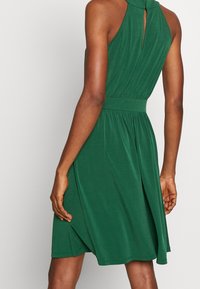 Femme portant une robe verte sans manches, à longueur genou, avec une taille cintrée et un détail dos en forme de trou de serrure, debout devant un fond uni.