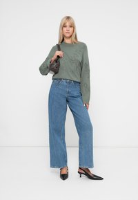 Pull vert olive clair avec broderie florale, jeans bleu taille haute et escarpins noirs à bout pointu. Petit sac à main sombre porté en bandoulière.