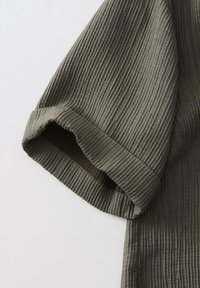 Tissu côtelé vert olive avec détail de manches courtes retroussées, présentant une texture subtile et une coupe décontractée.