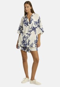 Modella che indossa un completo floreale crema e blu navy, composto da una camicia morbida con bottoni e pantaloni corti abbinati, abbinati a sneakers bianche.