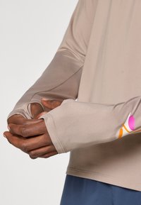 Cămașă sportivă cu mânecă lungă, de culoare bej, cu gauri pentru degete, având accente colorate cu logo pe mânecă și o textură de material neted și flexibil.