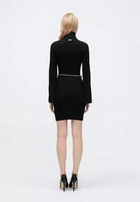 Fekete bordás kötött ruha magas gallérral, testhez simuló siluettel, hosszú ujjakkal és arany lánc övvel. Fekete magassarkú cipővel viselve.