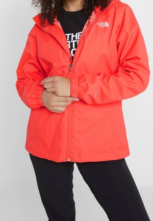 Personne portant une veste à capuche The North Face rouge vif avec un pantalon noir, mains jointes devant la zone de la fermeture éclair.