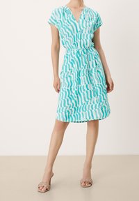 Robe turquoise avec un col en V, des manches courtes et une taille ceinturée. Elle présente un motif à rayures ondulées dans des nuances contrastées de bleu.