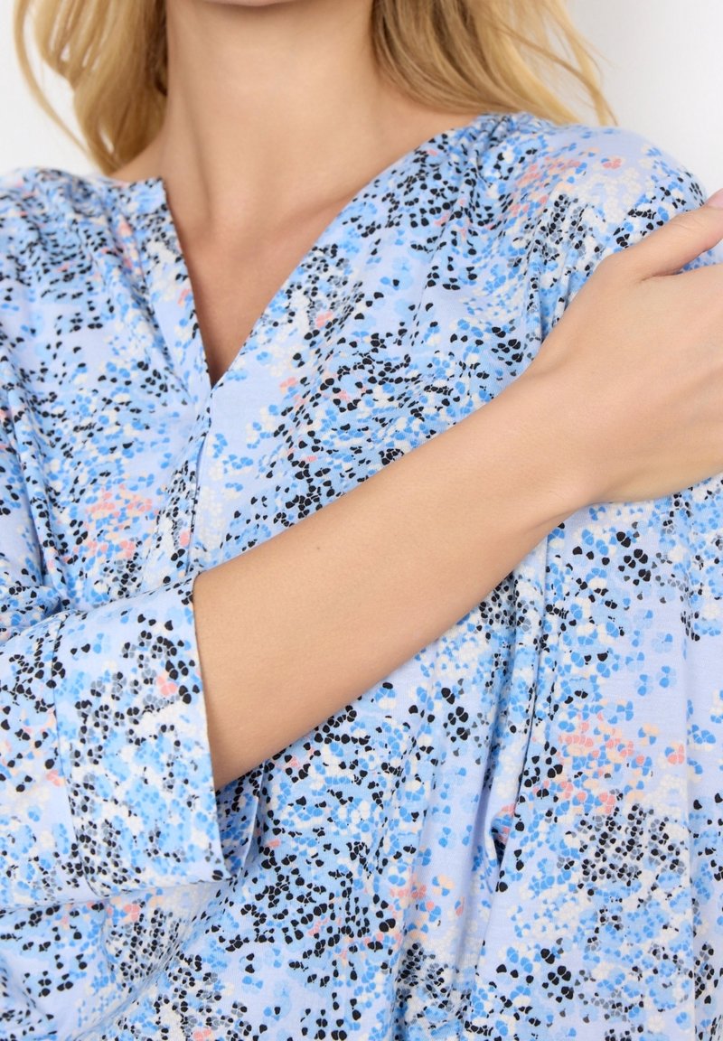Femme portant un chemisier bleu à motifs avec des taches noires, blanches et roses, sa main gauche reposant sur son épaule droite.
