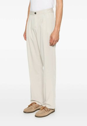 Uomo che indossa pantaloni beige plissettati e scarpe da barca marrone chiaro, in piedi davanti a uno sfondo bianco semplice.